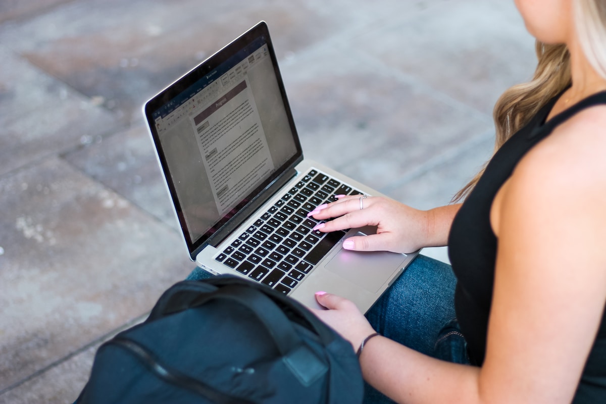 An athlete sitting outdoors with a backpack, typing on a laptop